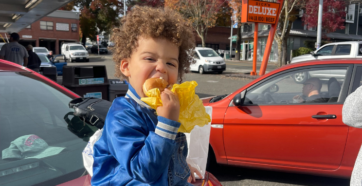 A small child sits on the trunk of a car eating a hamburger at Dick's Drive-In.