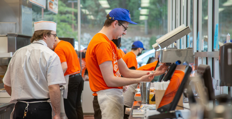 A group of restaurant crew members in orange and blue uniforms works behind the counter at Dick's Drive-In.