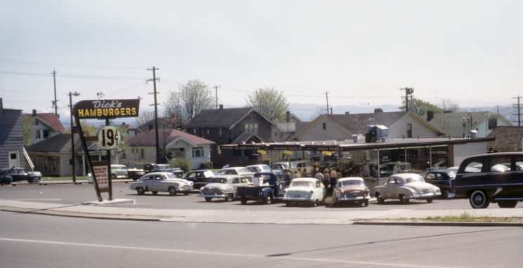 A historical photo of one of the original locations of Dick's Drive-In Restaurants, showing 1950s-era cars in the parking lot and a sign that reads, "Dick's Hamburgers, 19 cents."