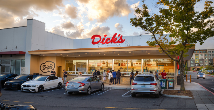 A view of a Dick's Drive-In location from the parking lot. A group of people gather in line to place orders under a brightly lit sign that reads, "Dick's."