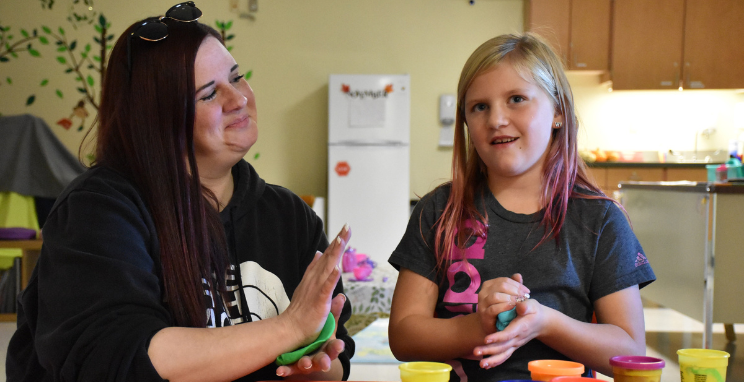 n adult looks at a child playing in a colorful classroom environment. Both are smiling, and there's a small kitchen in the background.